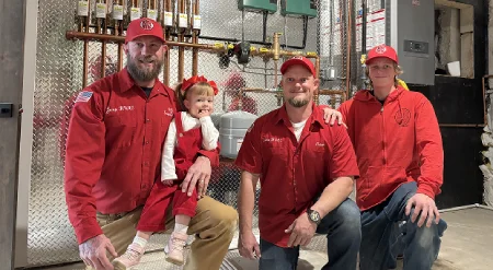 Group of three HVAC service technicians in red uniforms posing with a toddler girl in a mechanical room.