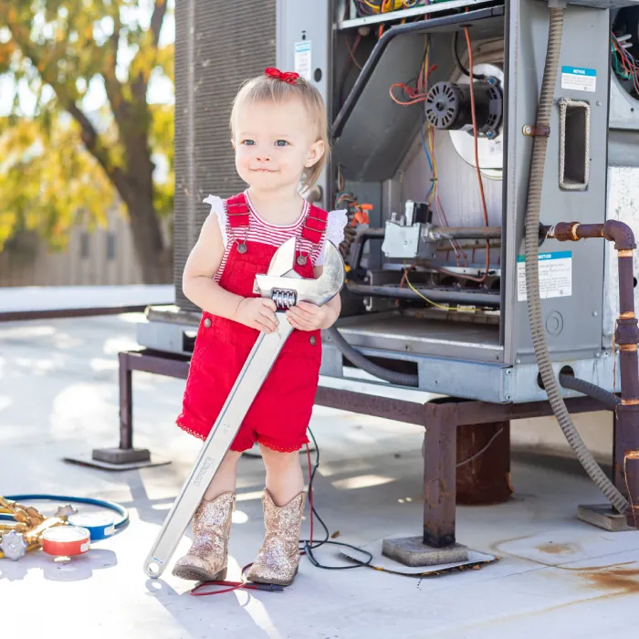 Toddler wearing red overalls holding a giant crescent wrench next to an open HVAC unit.