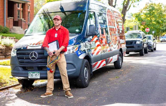 An HVAC technician wearing a red shirt and tool belt standing in front of a white residential garage door.