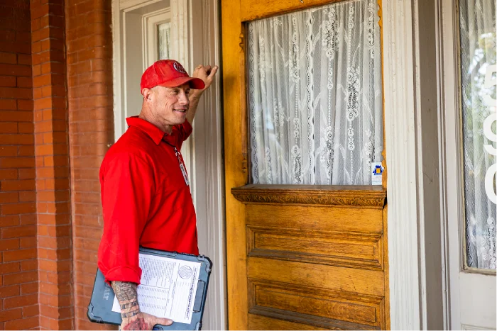 HVAC service technician in a red uniform knocking on a residential front door.
