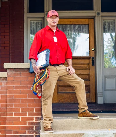 An HVAC technician holding service gauges standing on the porch steps of a residential home.