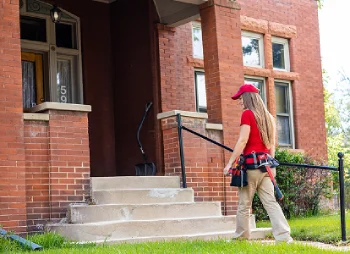 Side profile of a female HVAC technician wearing a tool belt walking up the concrete front steps of a brick residential home.