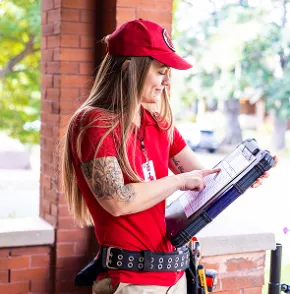 A female HVAC technician checking service details on a heavy-duty clipboard case outside a brick building.