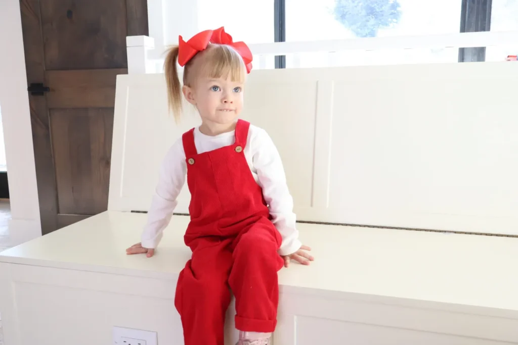 Toddler with a red hairbow and red overalls sitting on a white storage bench.