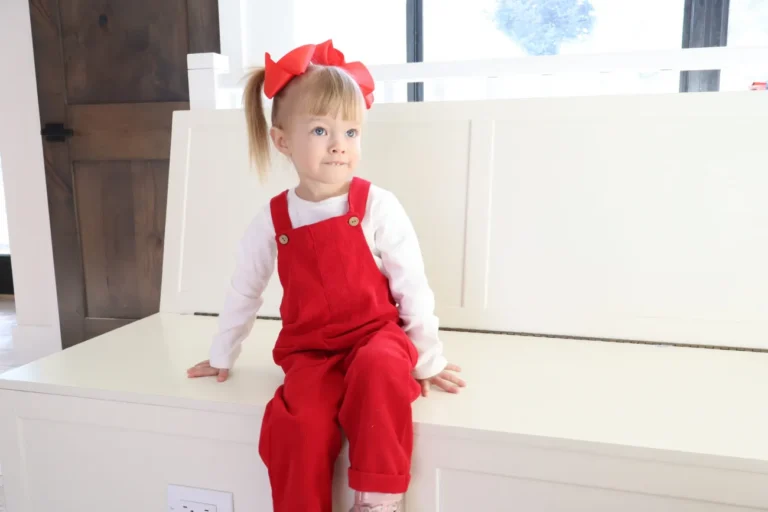 Toddler with a red hairbow and red overalls sitting on a white storage bench.