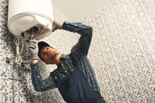 Plumber using a screwdriver to service an electrical panel under a wall-mounted water heater.