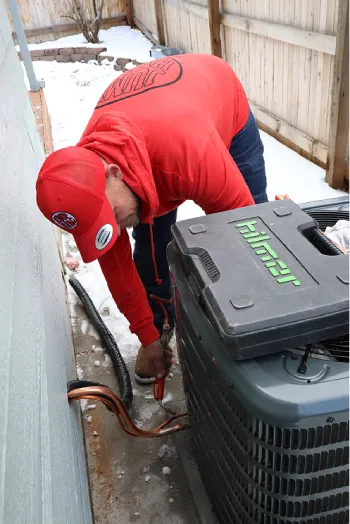 HVAC technician in a red hoodie and cap working on an outdoor AC unit in a snowy yard.